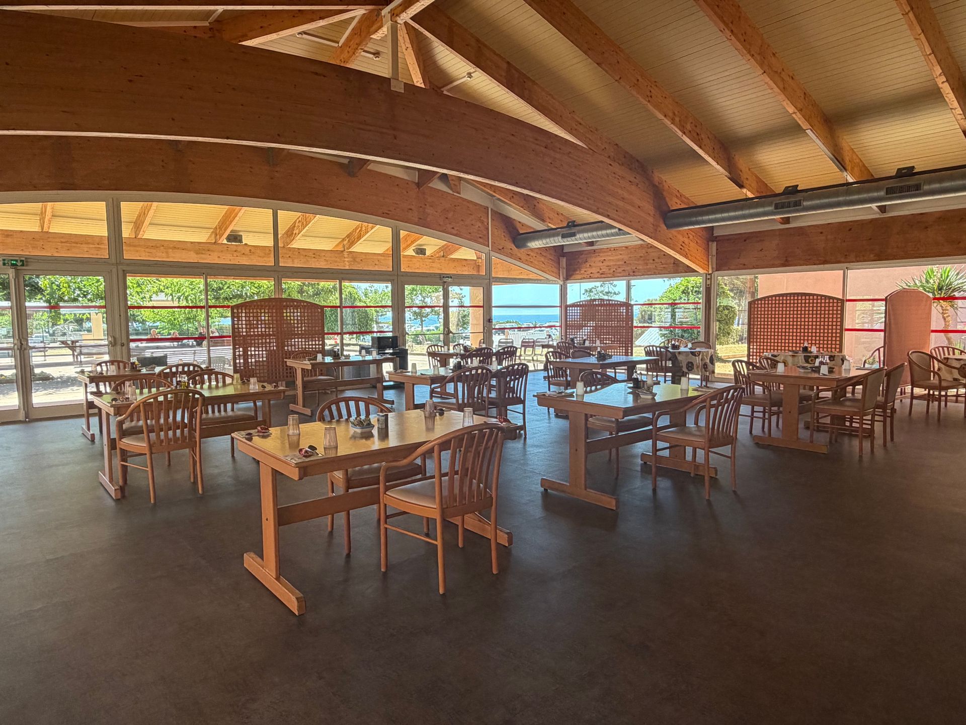 Salle à manger en bois avec tables et chaises sous un toit incurvé, donnant sur l'extérieur.