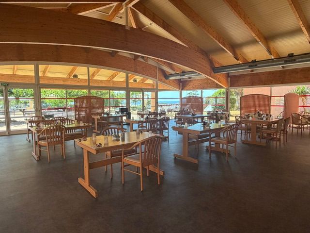 Salle à manger spacieuse avec poutres apparentes, tables, chaises et grandes fenêtres donnant sur l'extérieur.