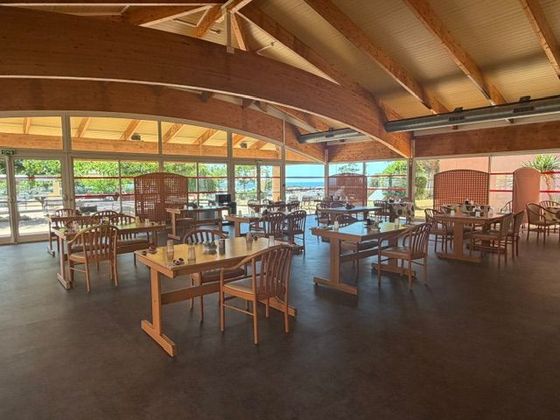 Spacieuse salle à manger en bois avec tables et chaises sous une charpente en bois courbée, ouverte sur la verdure extérieure.