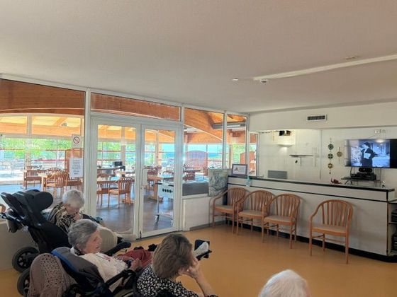 Des personnes assises dans un salon lumineux, face à une télévision et de grandes fenêtres sur le côté.