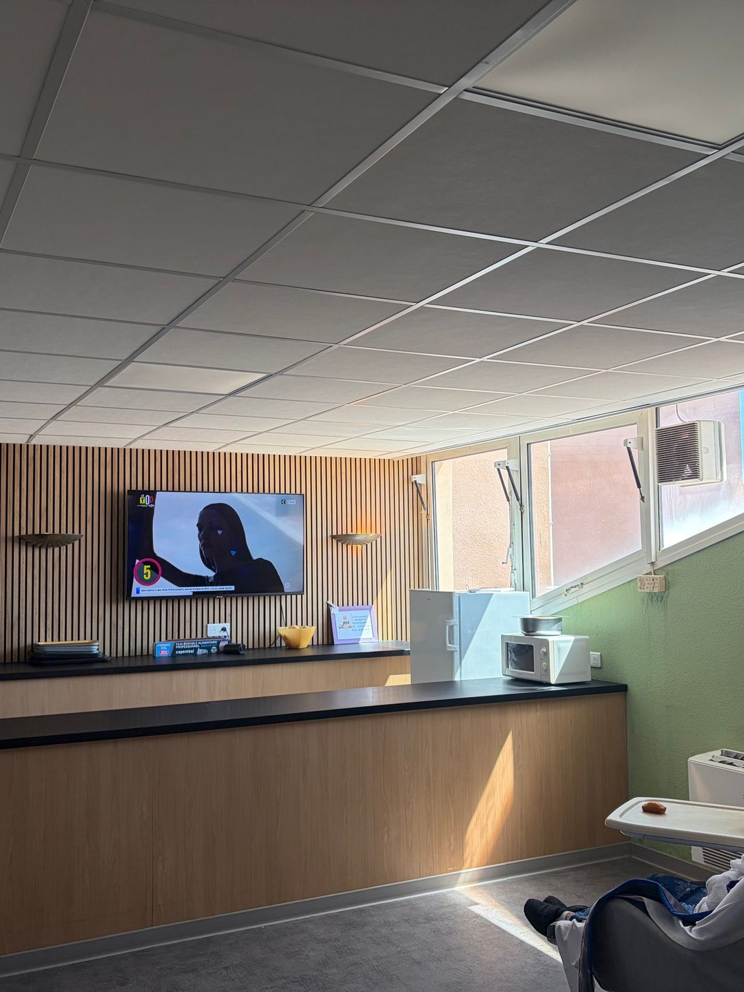 Salle commune moderne avec panneaux de bois, télévision murale et fenêtres baignées de lumière naturelle.