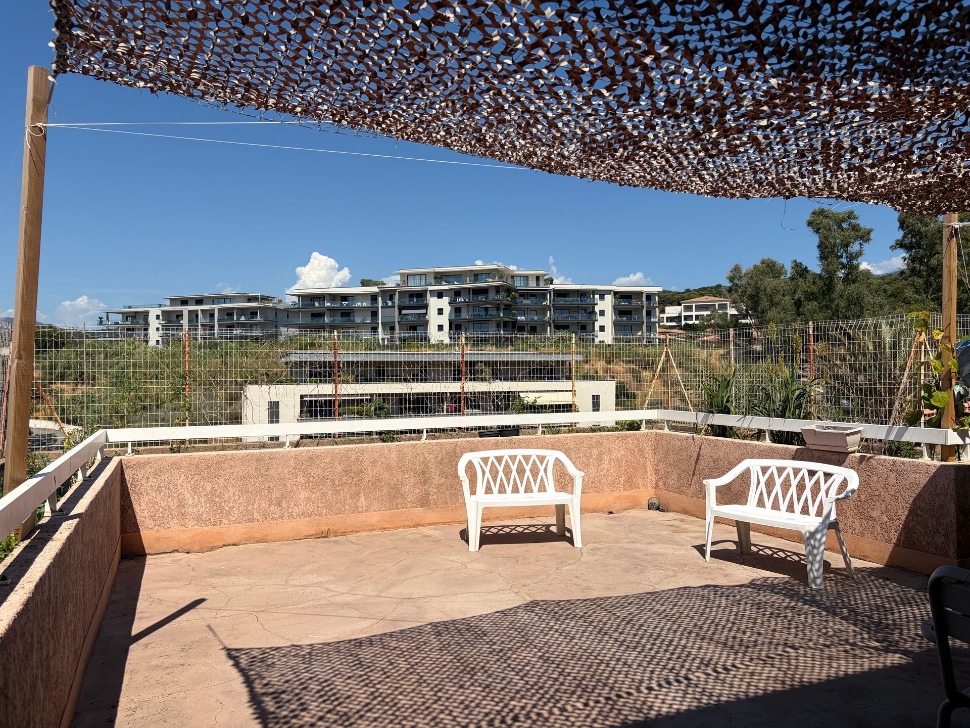 Terrasse ombragée avec deux chaises blanches donnant sur un bâtiment moderne et le ciel bleu.