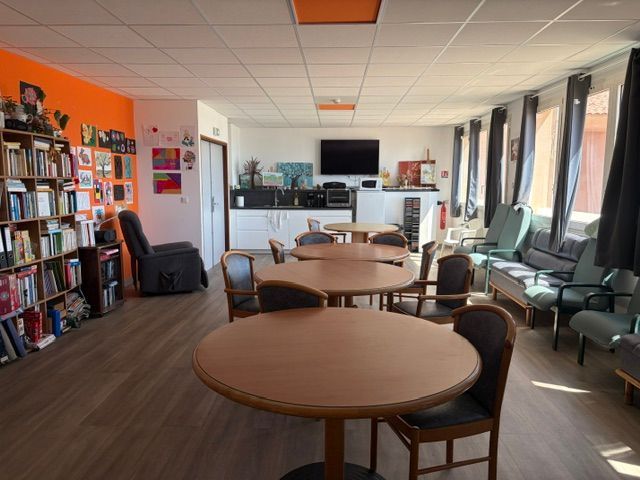 Salle commune lumineuse avec tables rondes, étagères, chaises et grandes fenêtres le long du mur de droite.