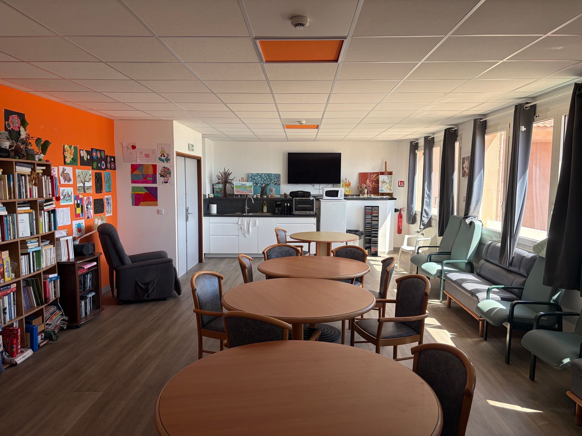Salle commune vide avec tables rondes, chaises, étagères et canapés près de grandes fenêtres.