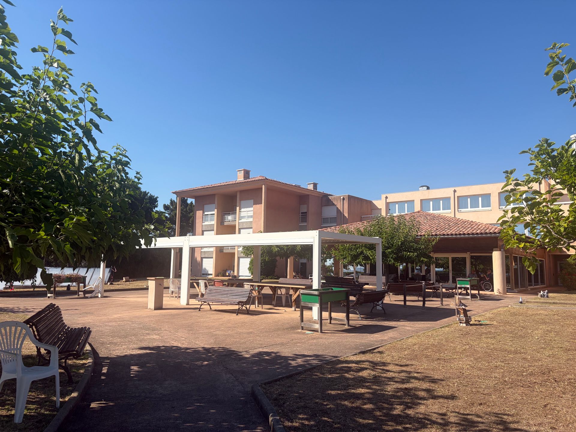 Terrasse extérieure avec tables, bancs et auvent blanc sous un ciel bleu clair, à côté d'un bâtiment à deux étages de l'EHPAD.