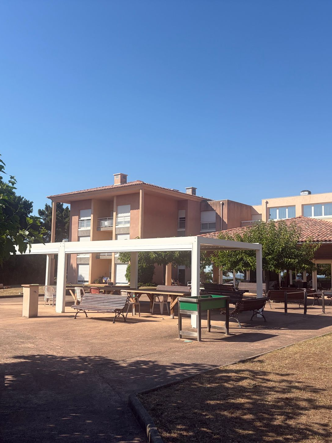 Terrasse extérieure avec tables de pique-nique sous une pergola blanche, à côté d'un bâtiment en briques rouges, par une journée ensoleillée.