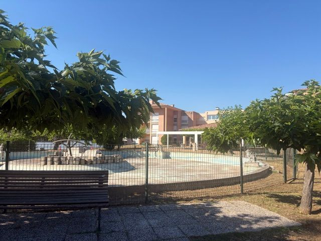 Cour ensoleillée avec un espace clôturé, des bancs, des arbres et un bâtiment en arrière-plan.