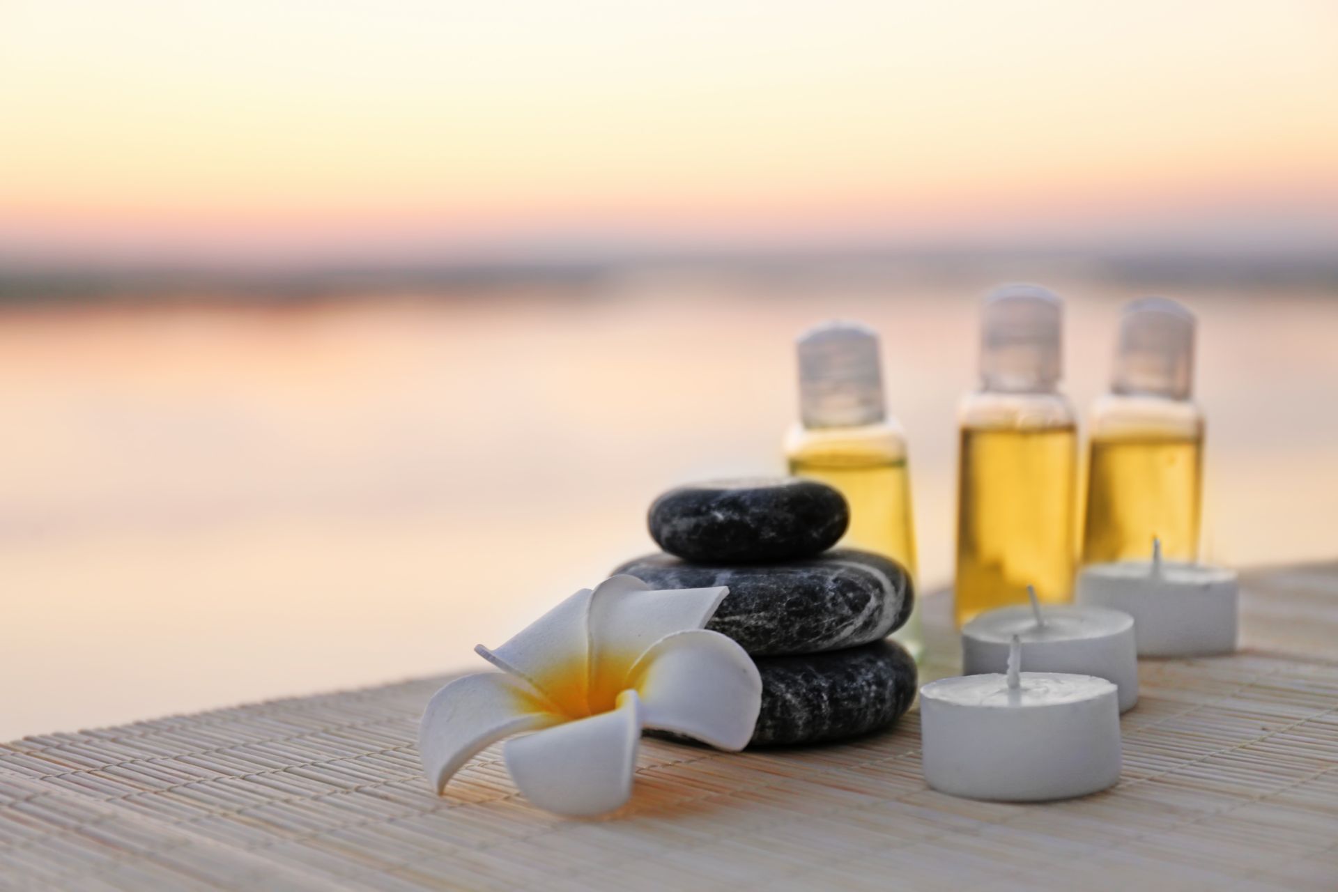 Ambiente de spa: piedras apiladas, aceites, velas y una flor de plumeria sobre una superficie de madera, con un fondo de atardecer borroso.