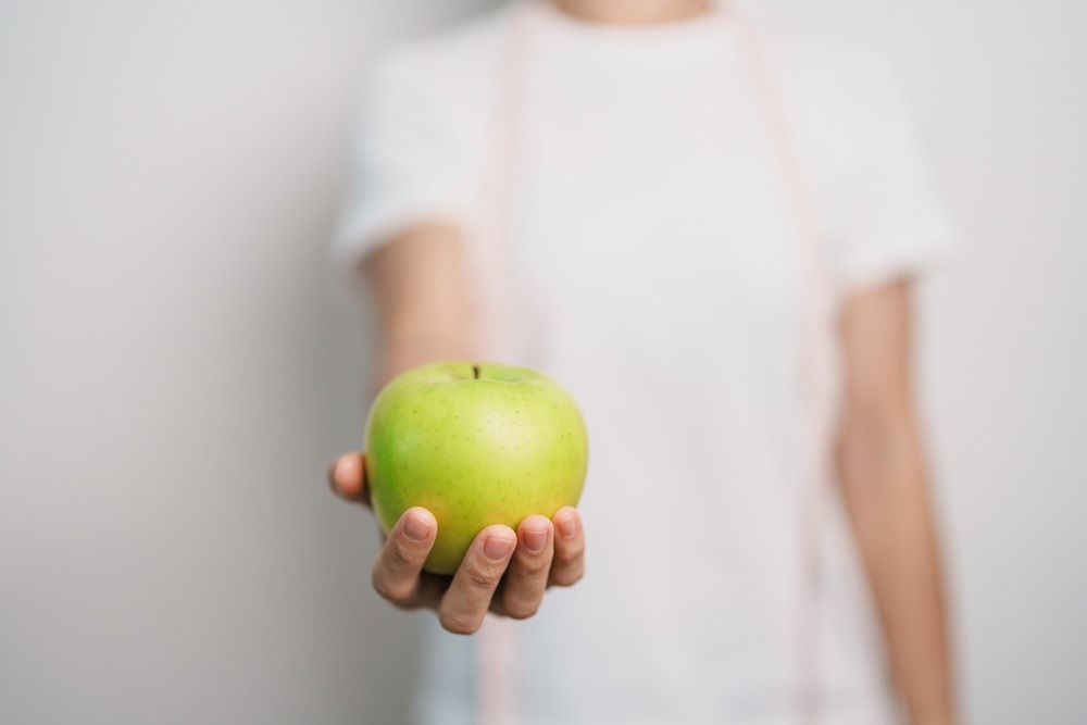 Persona sosteniendo una manzana verde. Camisa blanca sobre fondo blanco.