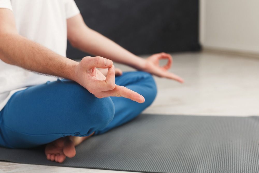 Persona en pose de meditación sobre una estera, con las manos en Gyan Mudra, vistiendo pantalones azules y camisa blanca.