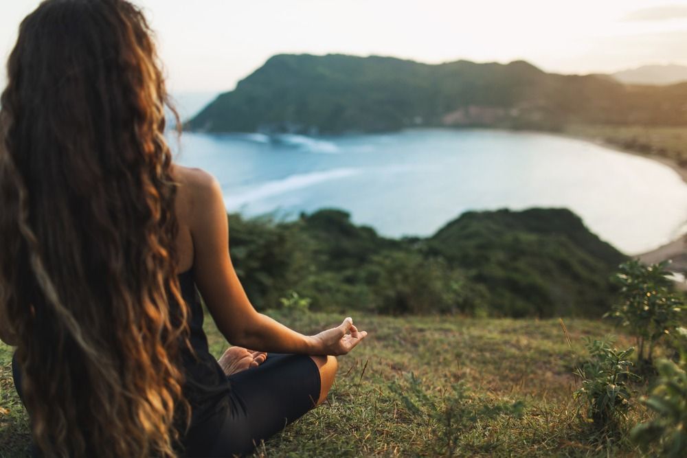 Mujer meditando en la cima de una colina con vistas a una bahía; luz de la hora dorada, cabello largo y castaño.