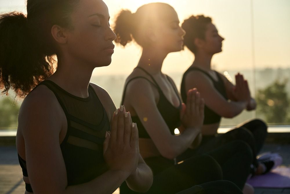 Tres mujeres meditando al aire libre al atardecer, con las manos juntas y los ojos cerrados.