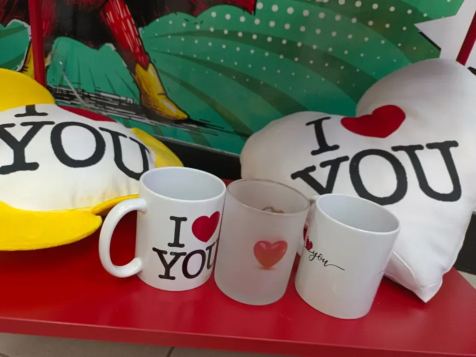 Tazas y cojines con motivos de corazones sobre una superficie roja, incluyendo artículos blancos con el texto 