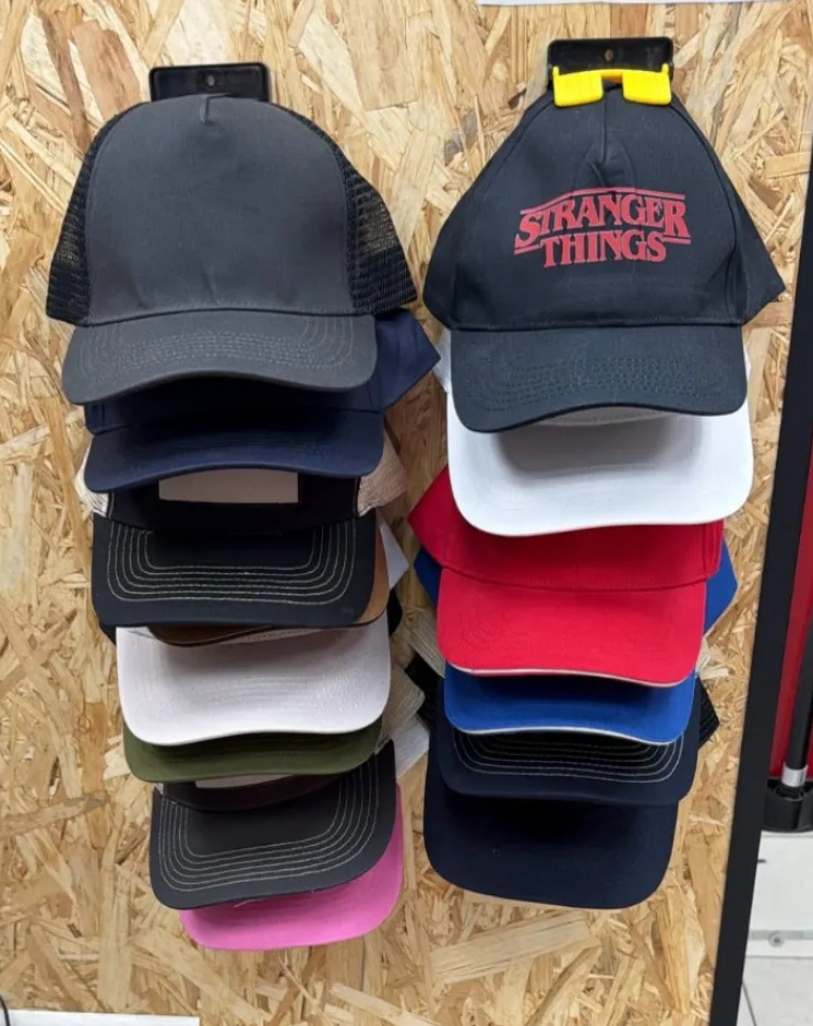 Gorras de béisbol apiladas en exhibición, incluyendo una gorra negra de Stranger Things y varias gorras de colores lisos.