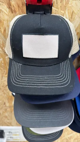 Pila de gorras de camionero de malla oscura con parches rectangulares blancos en la parte superior, expuestas en un estante.