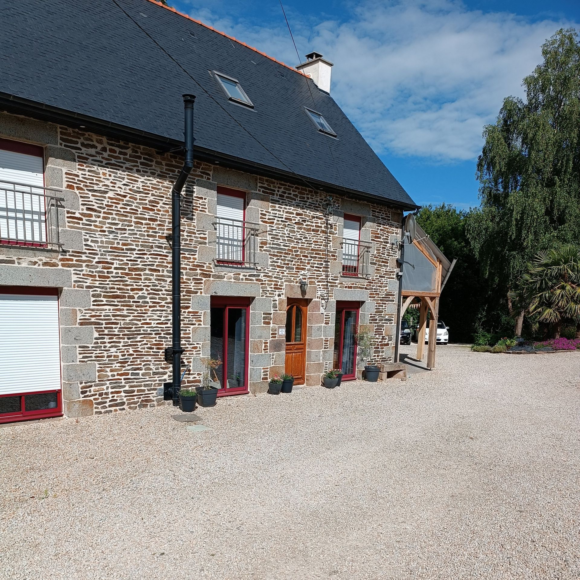 Maisonnette en pierre aux fenêtres et portes encadrées de rouge, donnant sur une cour de gravier sous un ciel bleu.