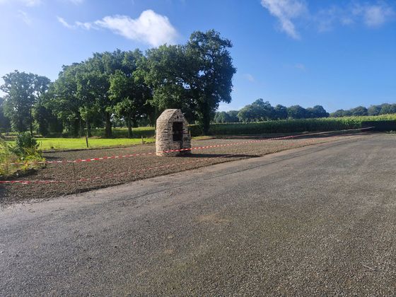 Route de campagne avec un petit autel de pierre à côté d'un champ de terre, des arbres verts et un ciel bleu