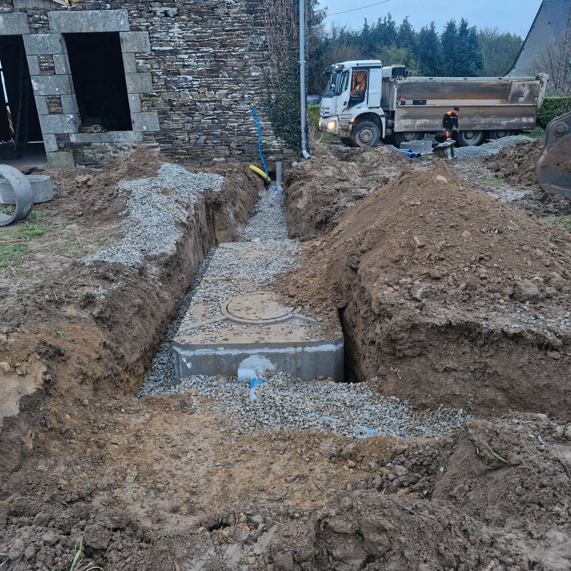 Excavation d'une tranchée à côté d'un bâtiment en pierre, avec des tuyaux en béton, du gravier et un camion-benne sur un chantier boueux.