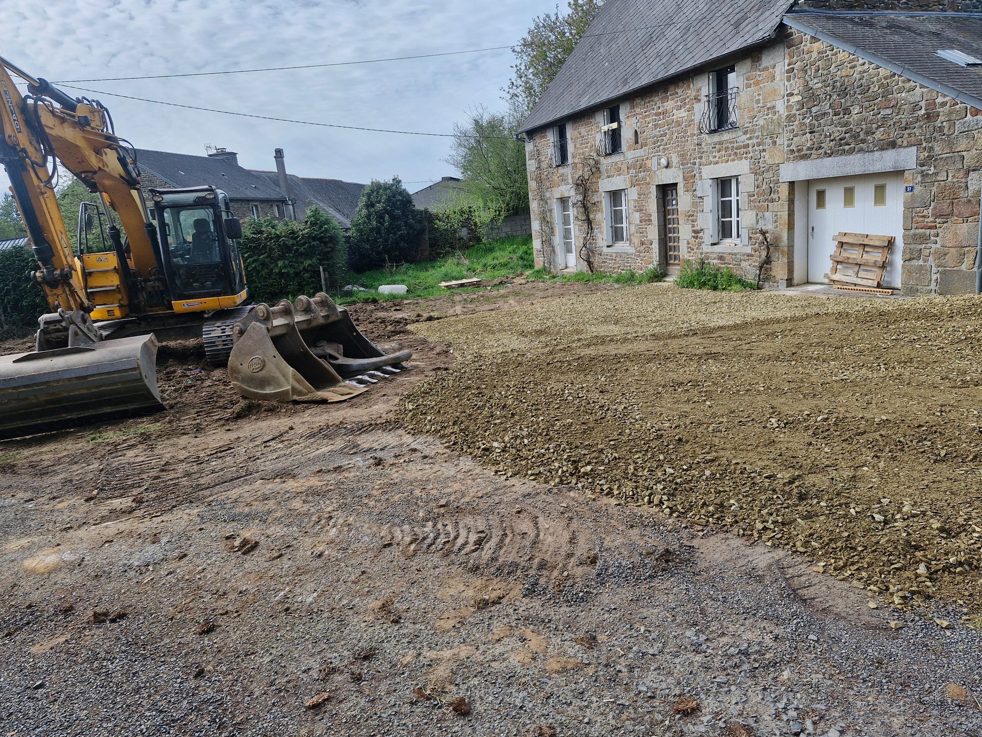 Chantier de construction avec une excavatrice répandant du gravier à côté d'une maison en pierre et d'une allée en terre.