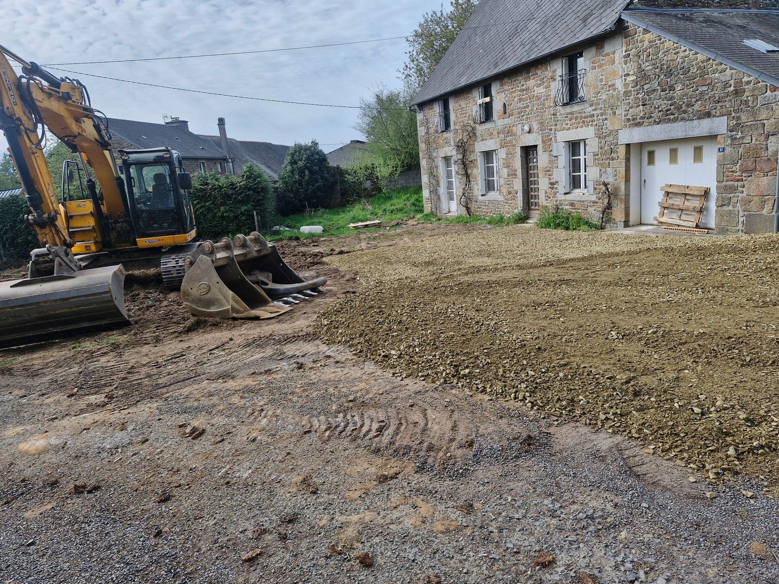 Chantier de construction avec une excavatrice nivelant du gravier à côté d'une maison et d'un garage en pierre.
