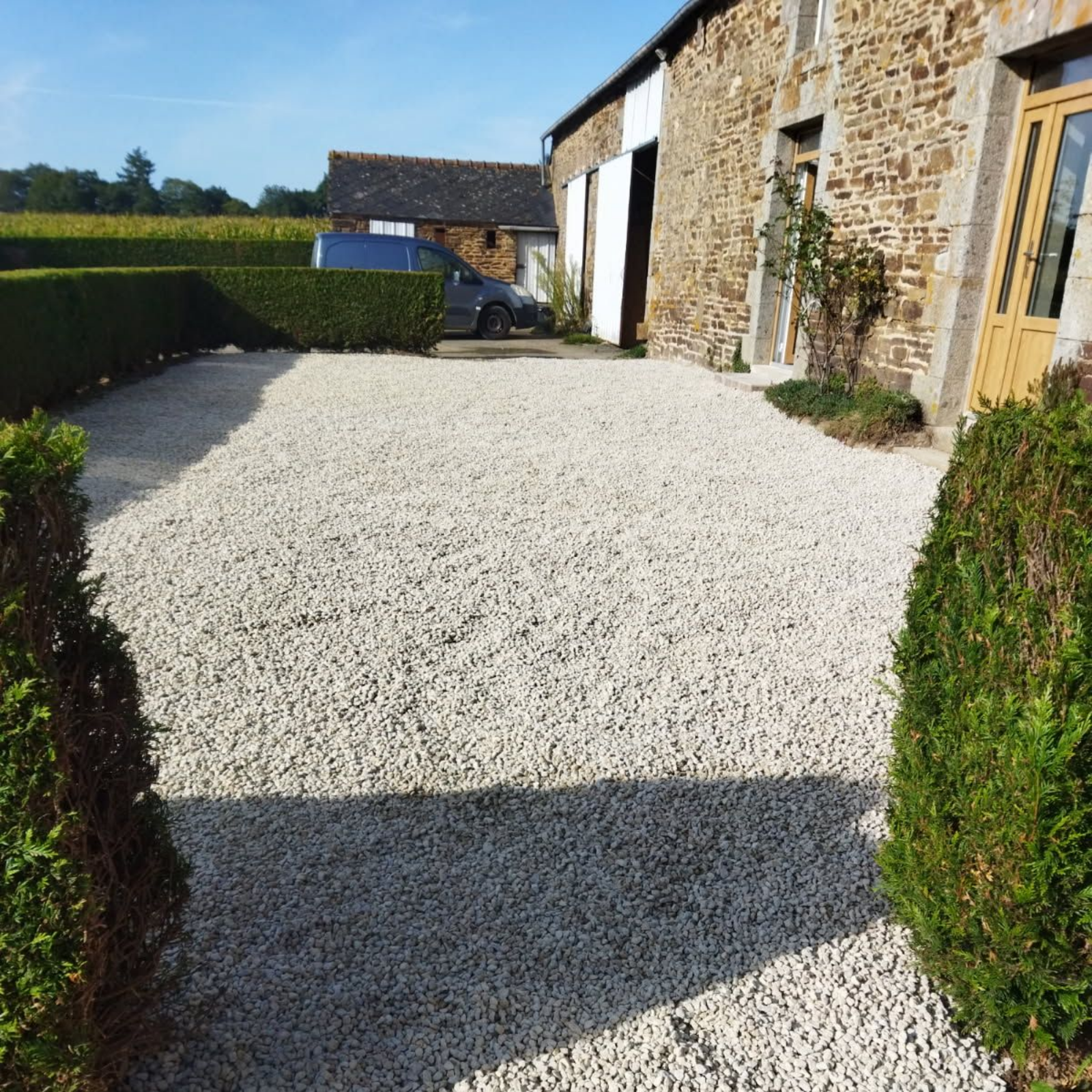 Allée de gravier longeant une maison en pierre et une haie, avec une cour ensoleillée et des dépendances en arrière-plan.