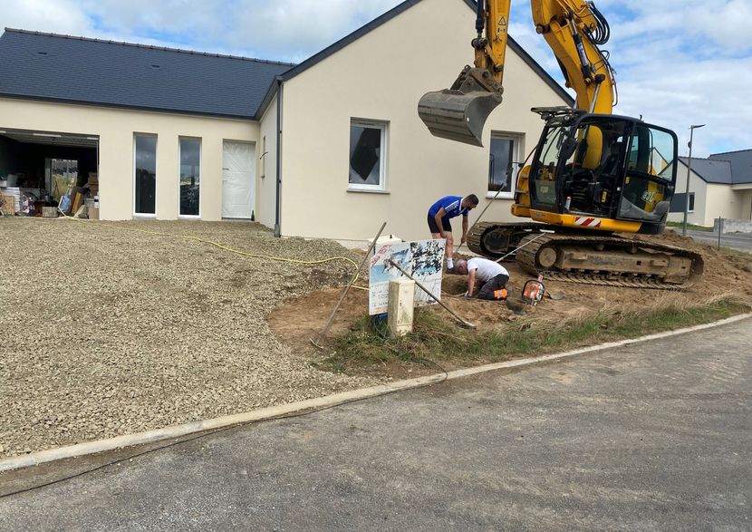 Un ouvrier installe une ligne électrique à côté d'une excavatrice près d'un bâtiment blanc et d'un terrain gravillonné.