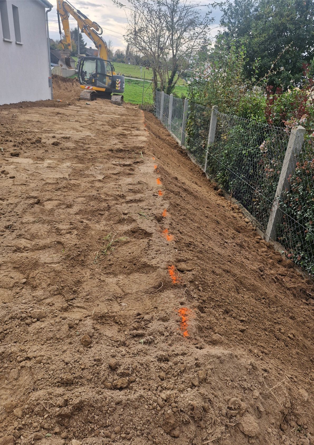 Terrain en terre fraîchement nivelé à côté d'une maison, avec des balises orange le long d'une clôture et des arbres sur la droite.