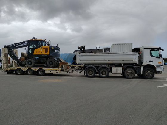 Un camion-benne blanc transporte de la machinerie lourde sur une remorque surbaissée dans un parking nuageux.
