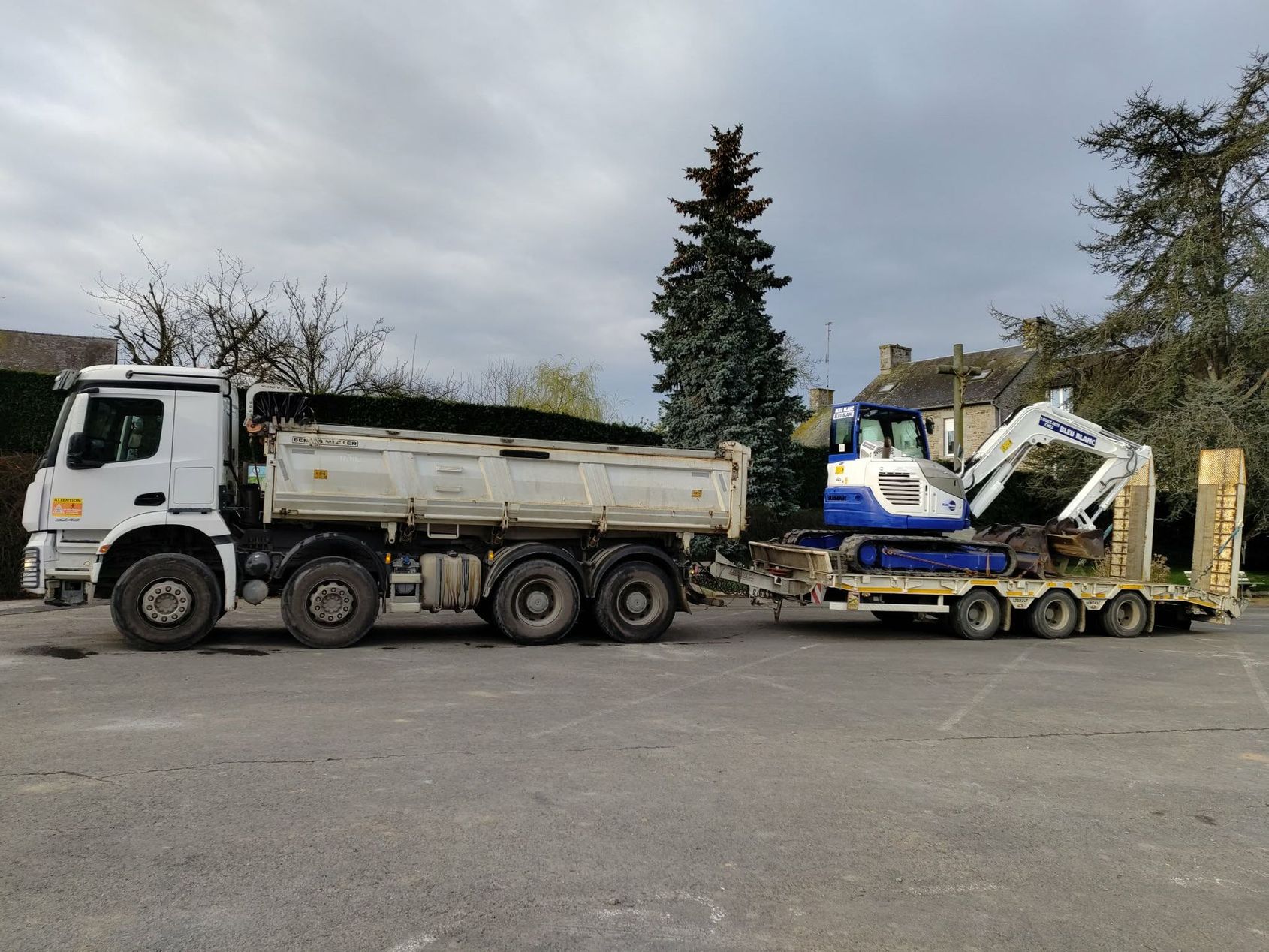 Un camion-benne et une pelle mécanique bleue sont chargés sur une remorque à plateau dans un parking.