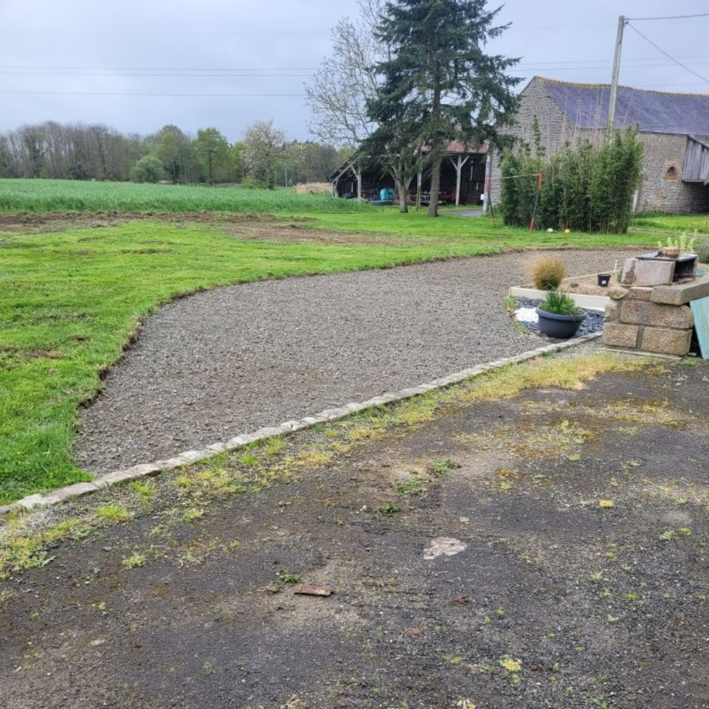 Chemin de gravier longeant une cour herbeuse, avec une jardinière en pierre et une ferme en arrière-plan.
