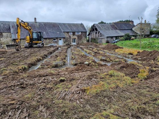 Une pelleteuse déblaye un champ boueux près de maisons en pierre.