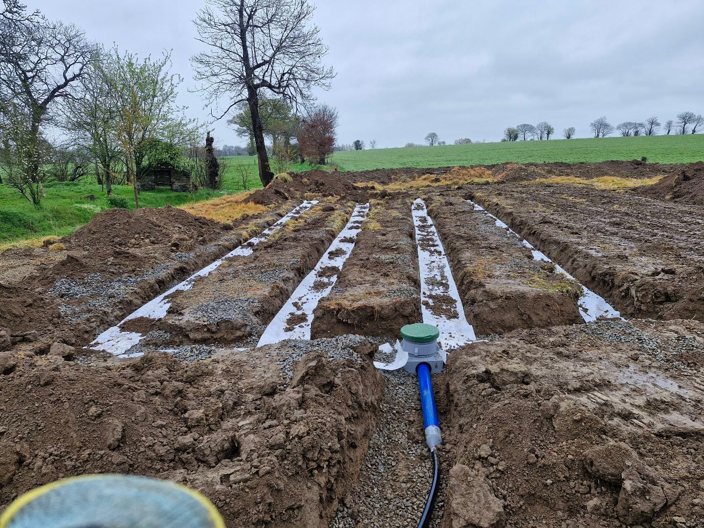 Champ labouré avec des tranchées boueuses et des tuyaux blancs, sous un ciel nuageux avec des arbres en arrière-plan