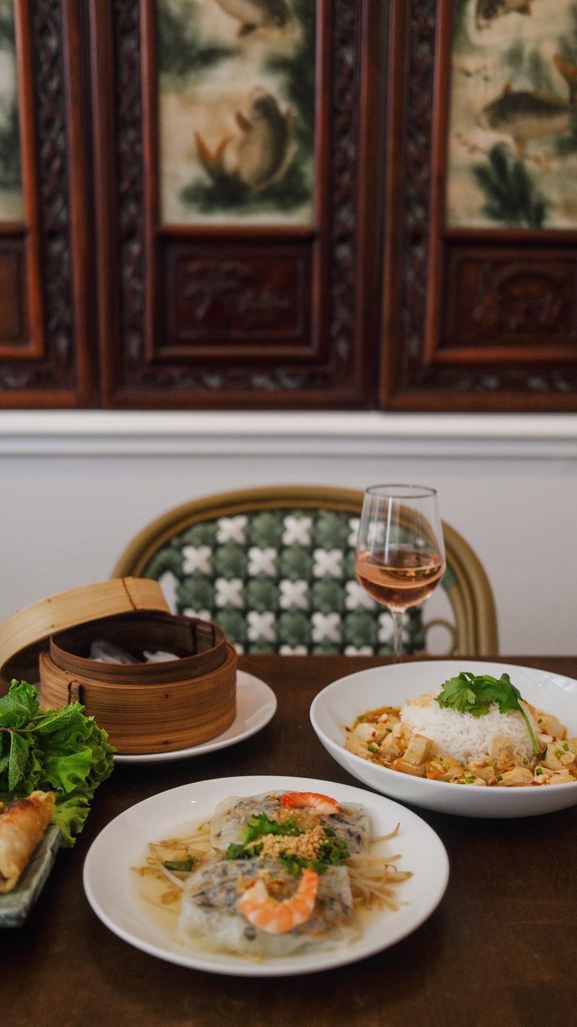 Table de restaurant à l'éclairage tamisé avec des pâtes servies à l'assiette, un panier vapeur en bambou et un cocktail à côté de chaises à motifs.
