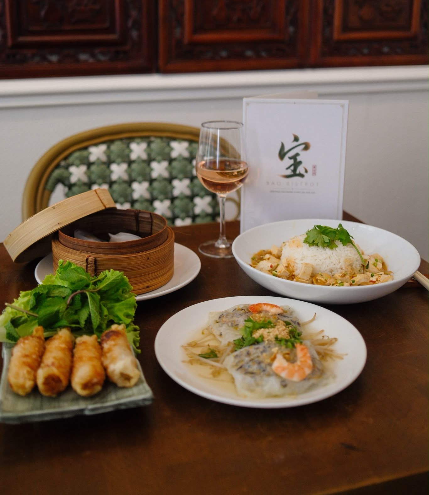 Un repas chinois est servi sur une table sombre, avec des petits pains, des rouleaux de printemps, des nouilles, de la soupe, du thé et un panier tressé.