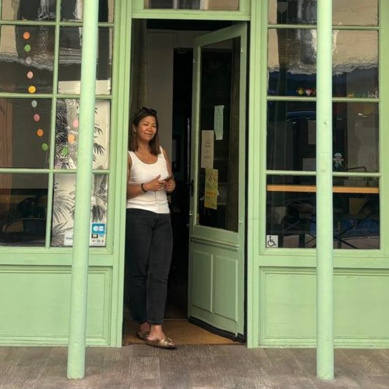 Une personne se tient debout dans l'embrasure d'une porte de magasin verte, tenant la porte ouverte et souriante.