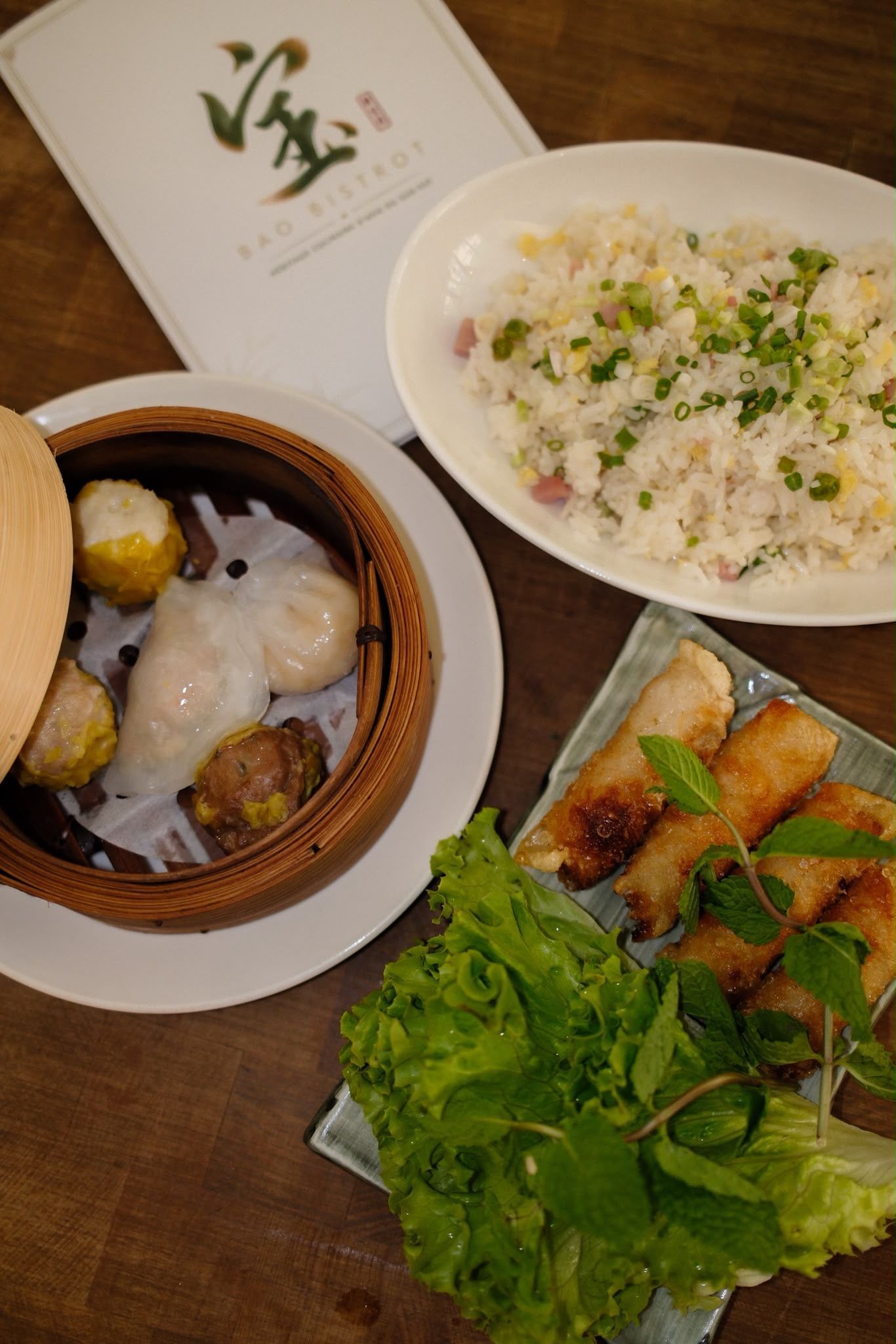 Assortiment de plats asiatiques sur une table en bois, comprenant du riz, des raviolis, des rouleaux de printemps et des légumes verts.