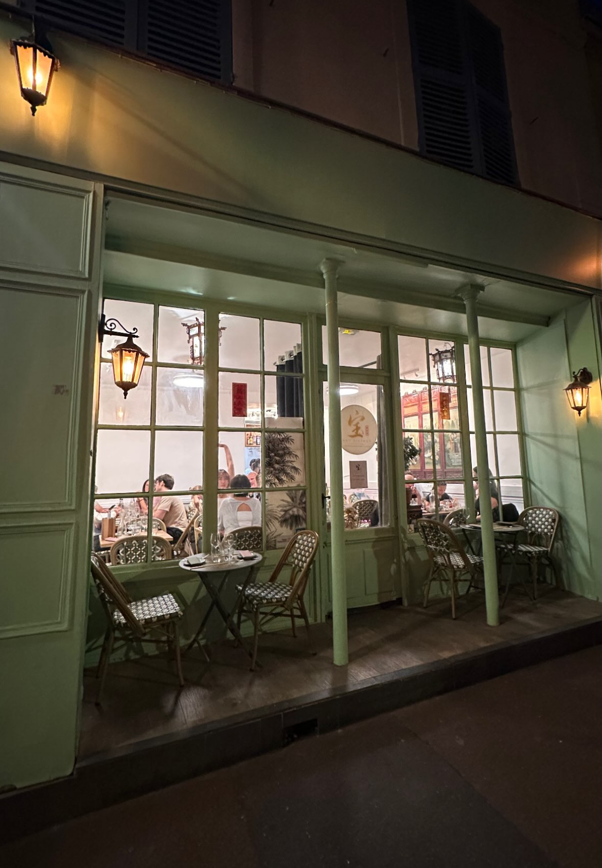 Vue nocturne d'un café chaleureux aux fenêtres encadrées de vert et à la terrasse illuminée par une douce lumière.