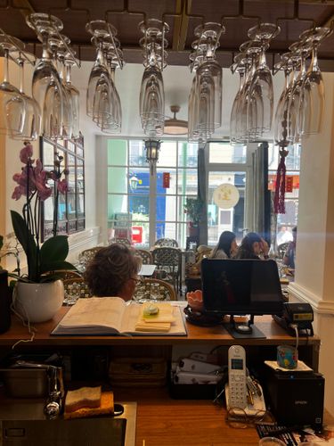 Intérieur d'un café avec des verres à vin suspendus, un comptoir, un livre ouvert et des personnes assises près de la fenêtre.
