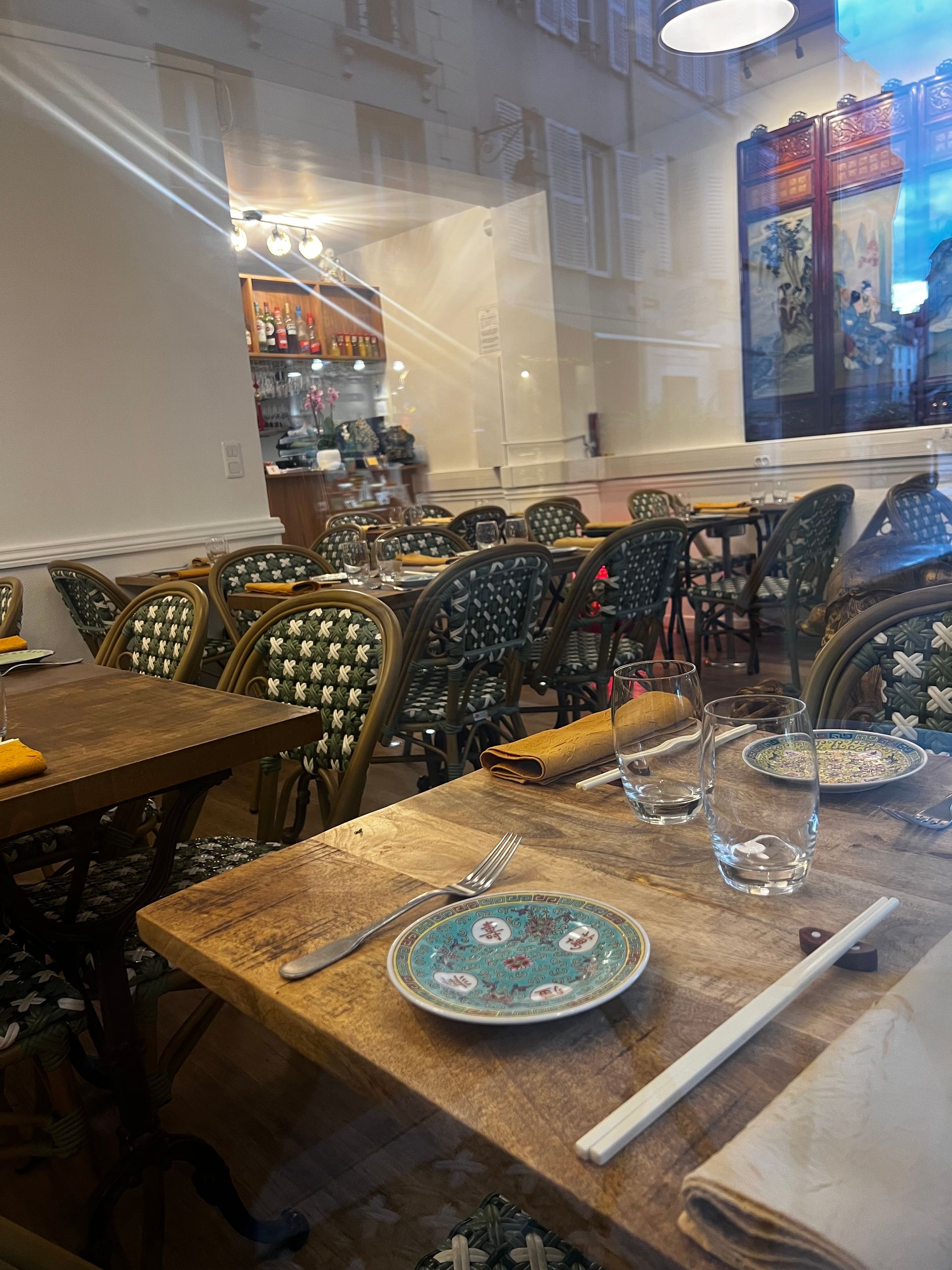 Salle à manger de café avec chaises à motifs, tables en bois dressées avec assiettes et verres, et grandes fenêtres.