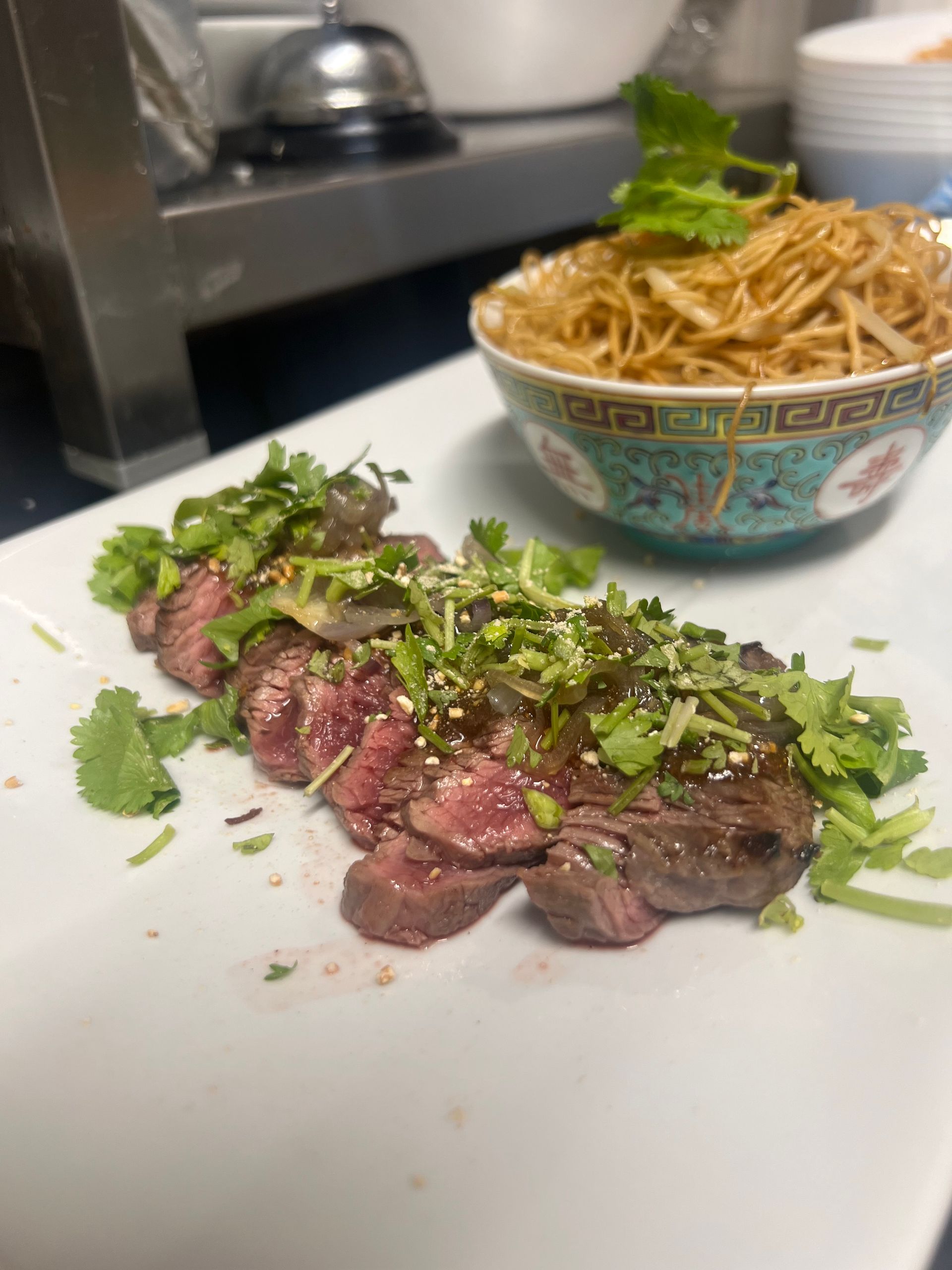 Steak tranché garni d'herbes sur une assiette blanche à côté d'un bol de nouilles dans une cuisine