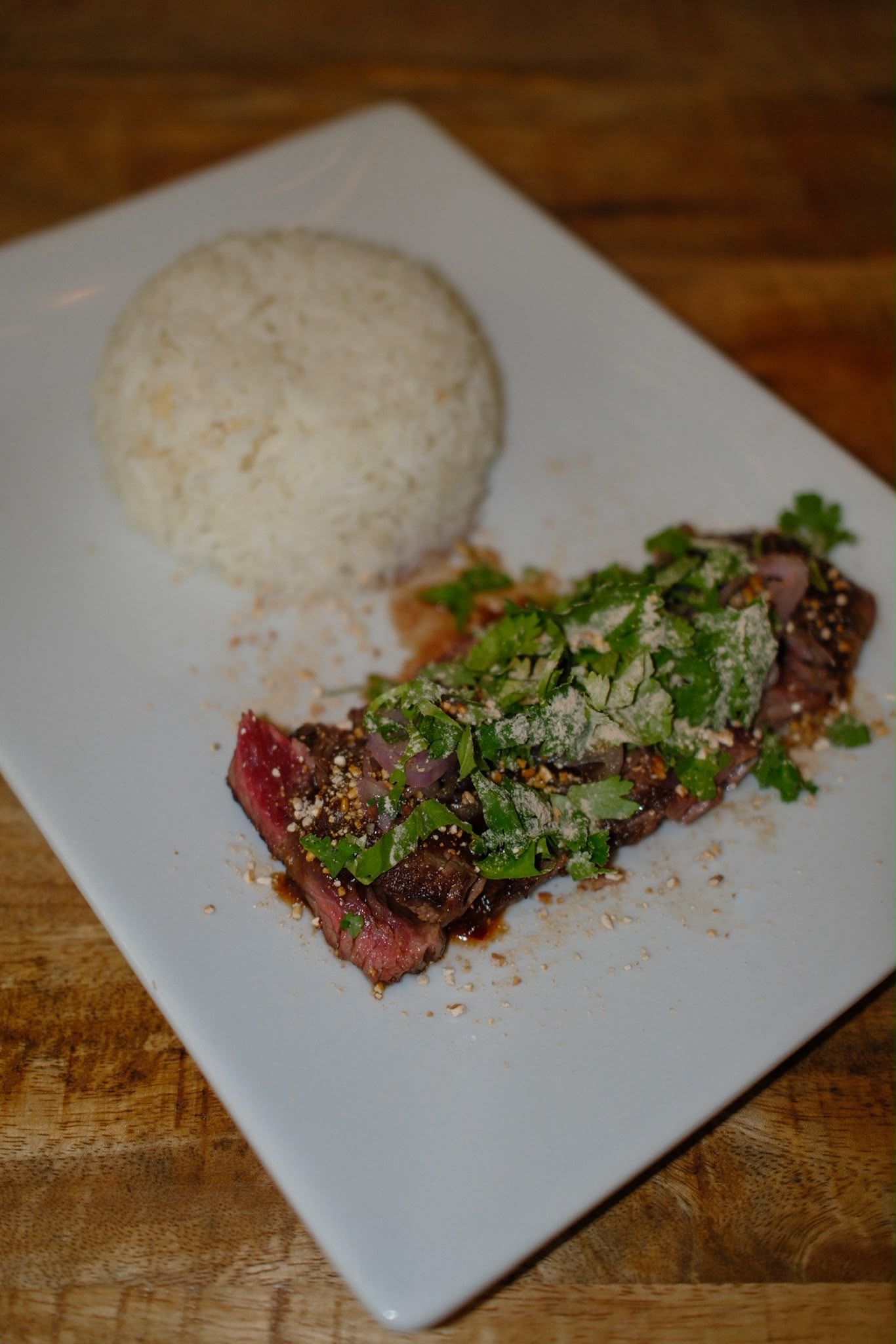 Riz blanc avec de la viande grillée garnie d'herbes hachées, servi sur une assiette blanche rectangulaire posée sur une table en bois.