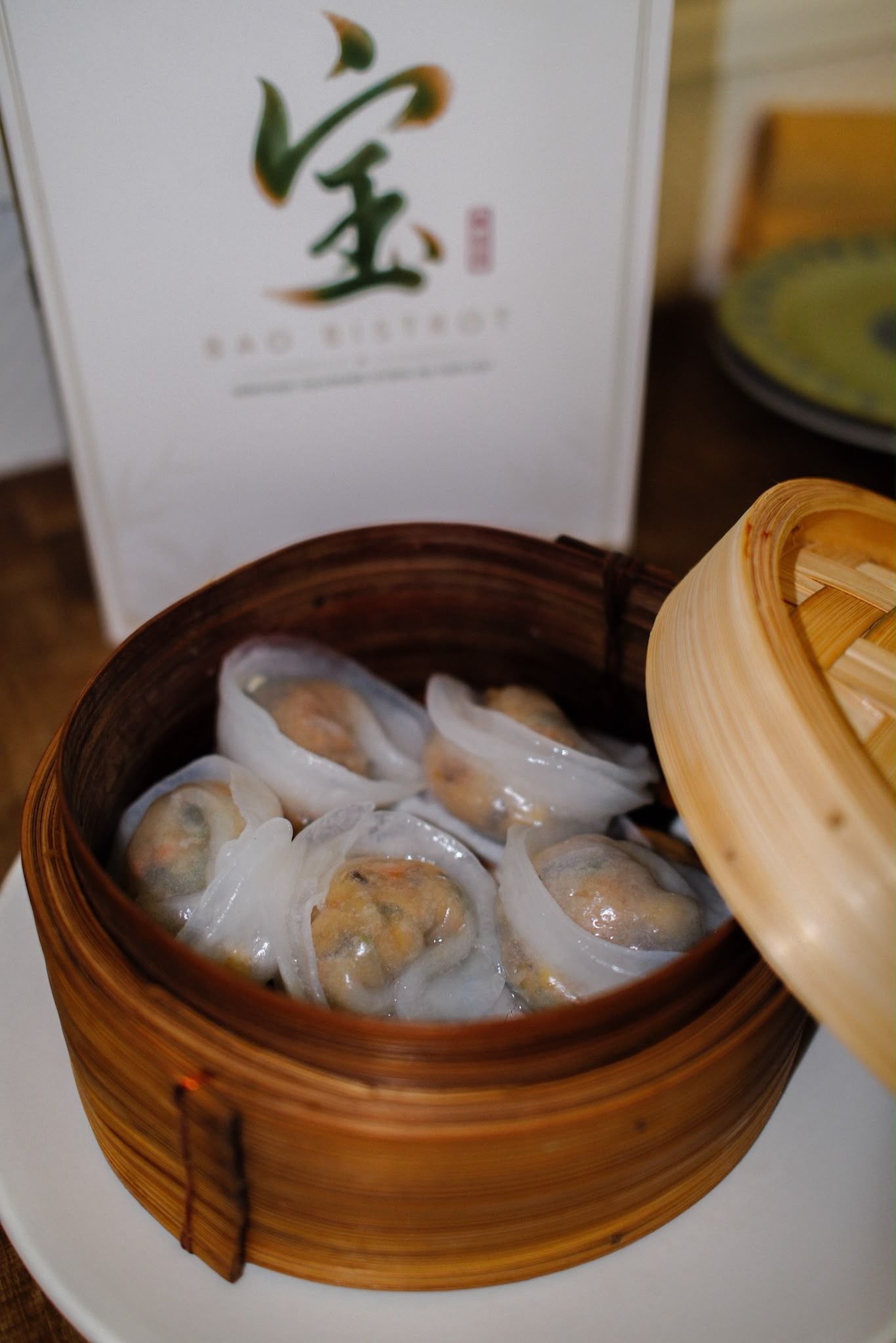 Panier vapeur en bambou contenant des raviolis assortis sur une assiette blanche, avec un menu de restaurant en arrière-plan.