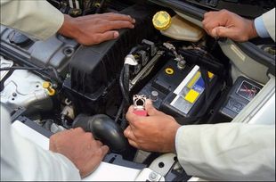Manos trabajando en el motor de un coche, con la batería y las herramientas visibles en el compartimento del motor.