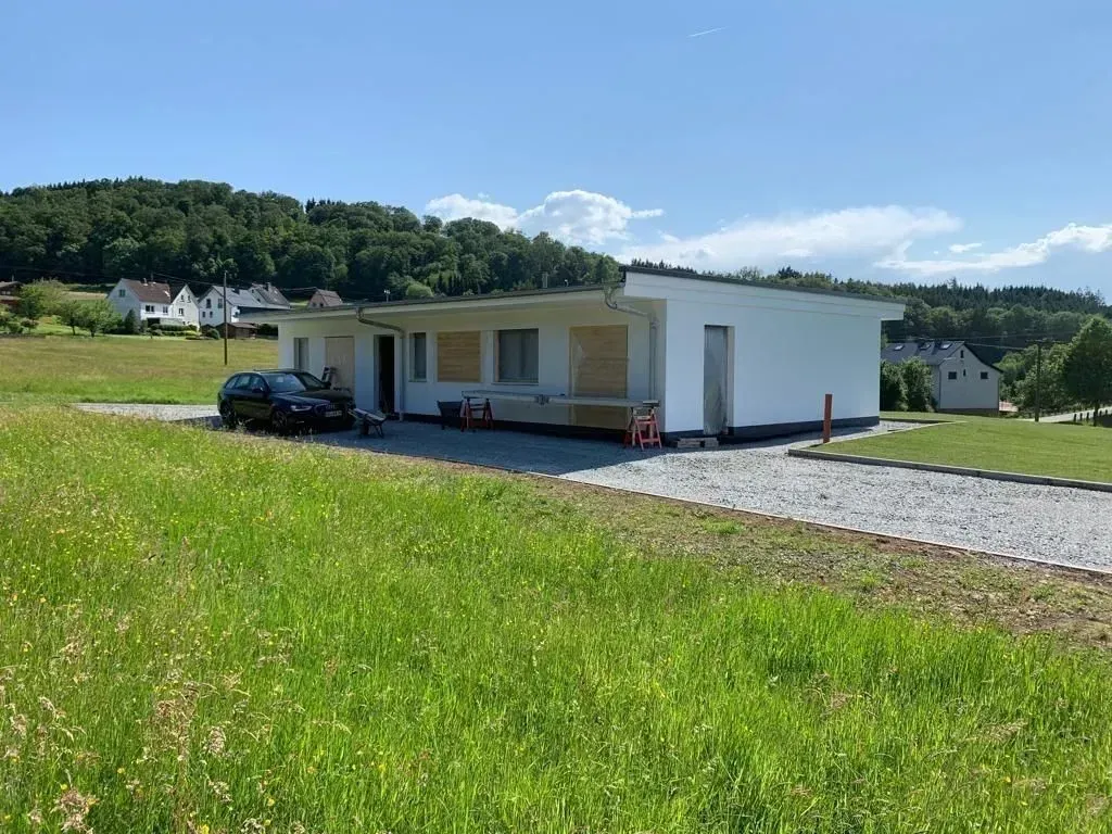 Modernes weißes Haus mit davor geparktem Auto, eingebettet in eine grasbewachsene ländliche Landschaft unter blauem Himmel