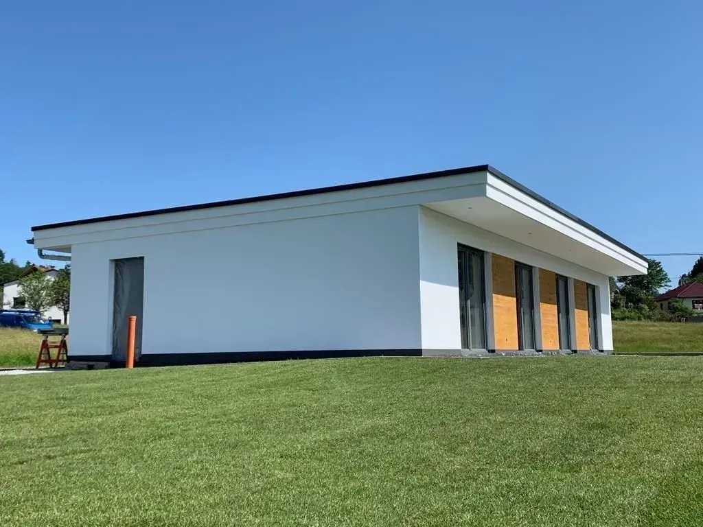 Weißes, modernes Gebäude mit schrägem Dach auf einem grasbewachsenen Hügel unter klarem, blauem Himmel