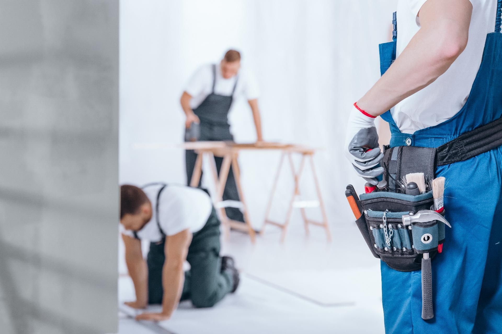 Arbeiter montieren einen Tisch in einem hellen Raum; im Vordergrund ist ein Werkzeuggürtel zu sehen.