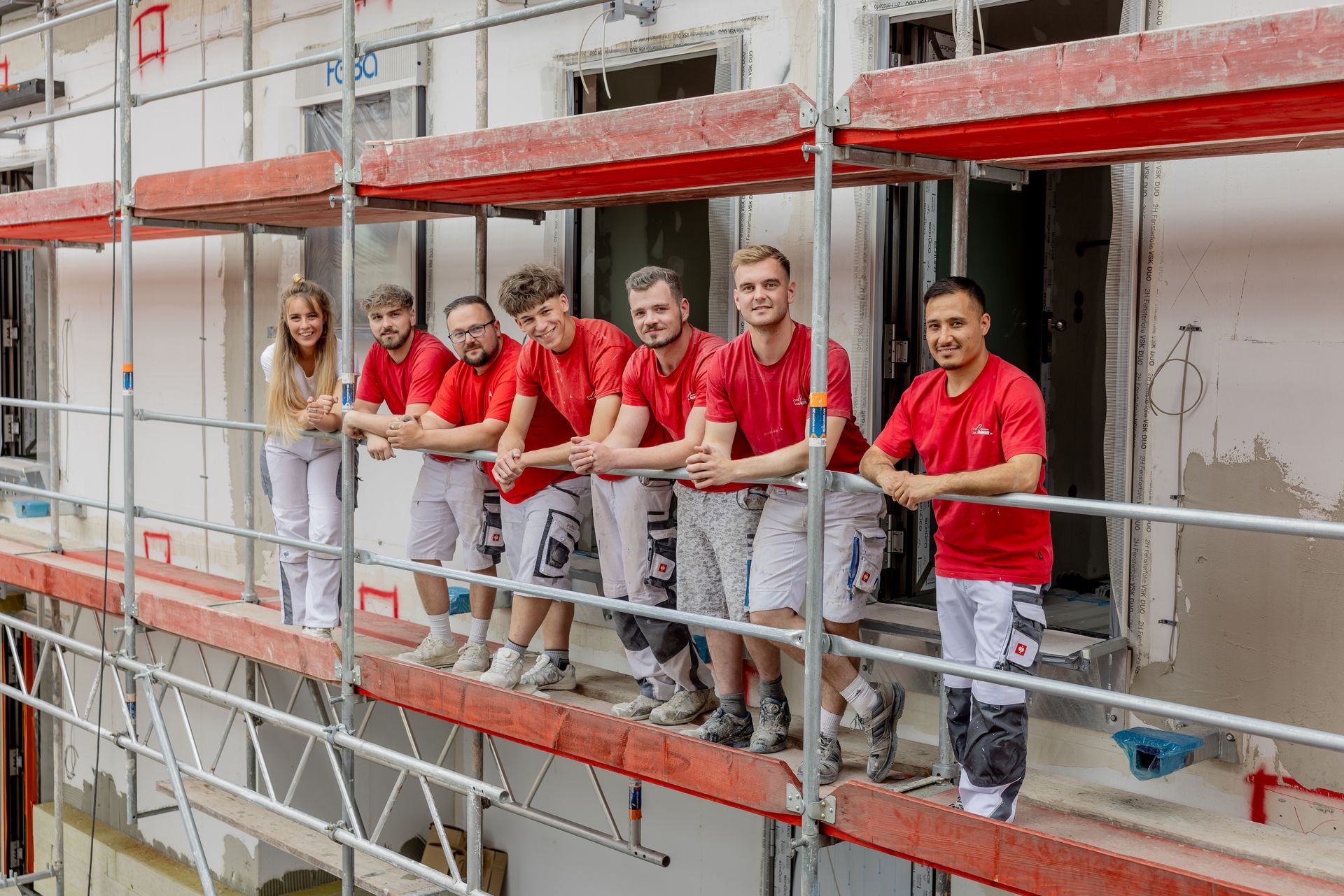 Zwei Arbeiter in roten Hemden halten ein Stück weiße Tapete an eine kahle Wand und messen es aus.