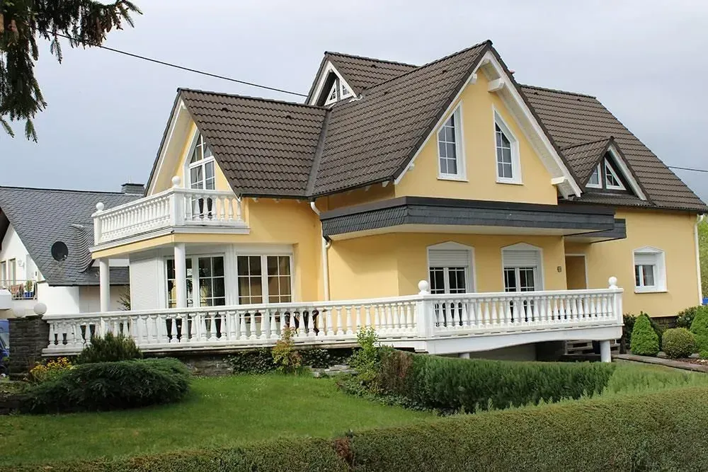 Gelbes Haus mit weißen Zierleisten und umlaufender Veranda in einem grünen Garten