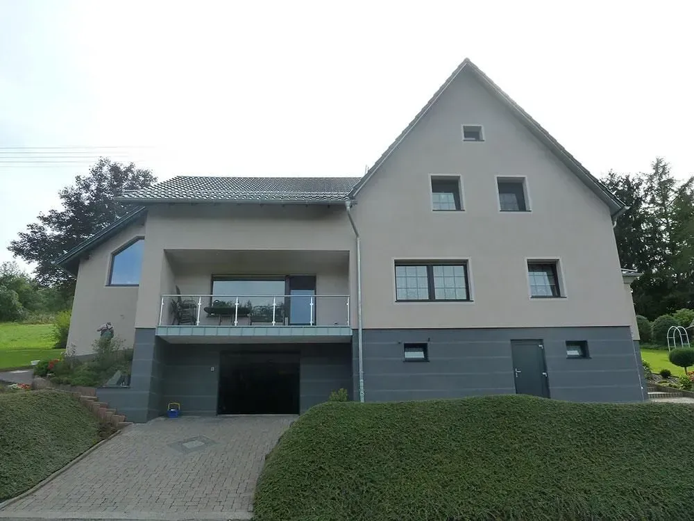 Modernes graues Haus mit Giebeldach, Balkon, Garage und schräger Auffahrt, umgeben von Grün