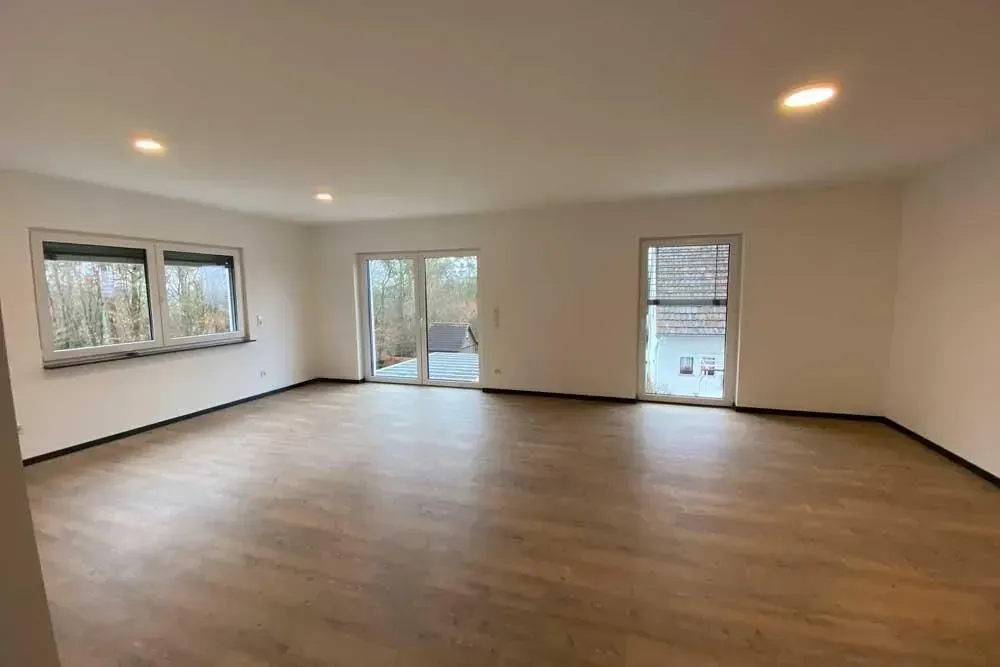 Leeres beigefarbenes Wohnzimmer mit Holzboden, Einbaustrahlern und Glasschiebetüren.